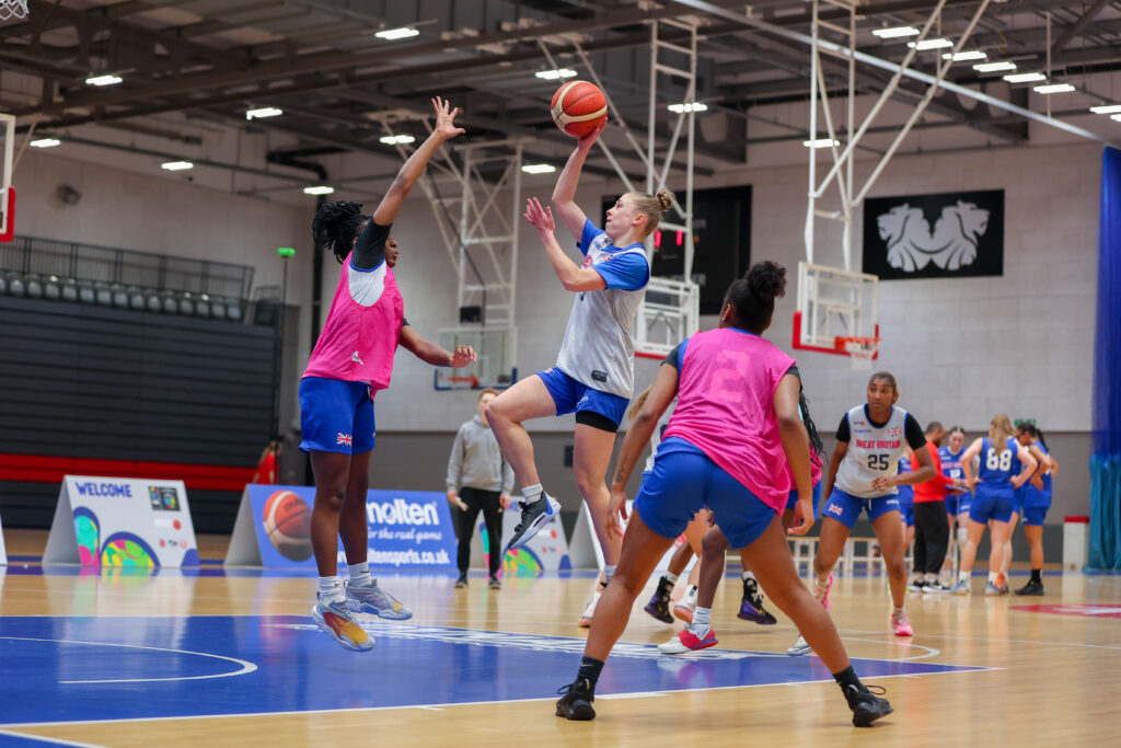 Young women playing basketball. One young woman has the ball and a second player wearing a pink bib is defending.