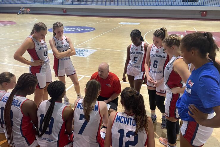 Donnie MacDonald is knelt by a basketball court. He is wearing a red polo shirt. He is surrounded by the GB under 16 women's basketball team. They are wearing mainly white kit.