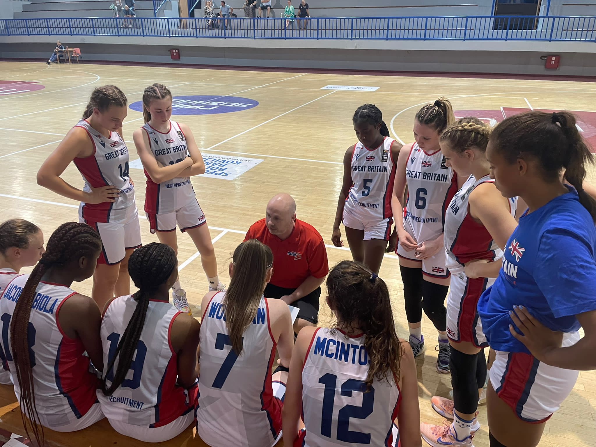 Donnie MacDonald is knelt by a basketball court. He is wearing a red polo shirt. He is surrounded by the GB under 16 women's basketball team. They are wearing mainly white kit. 